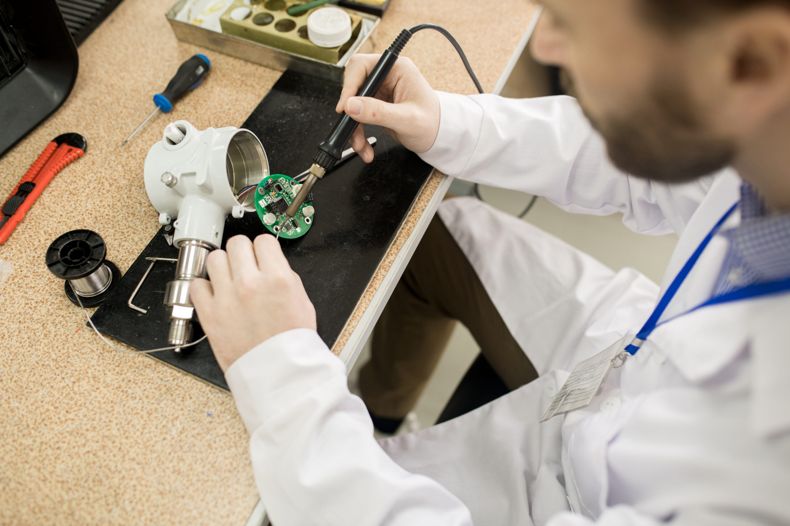 Close-up of busy male engineer in lab coat sitting at table with tools and soldering pressure transducer motherboard while assembling device