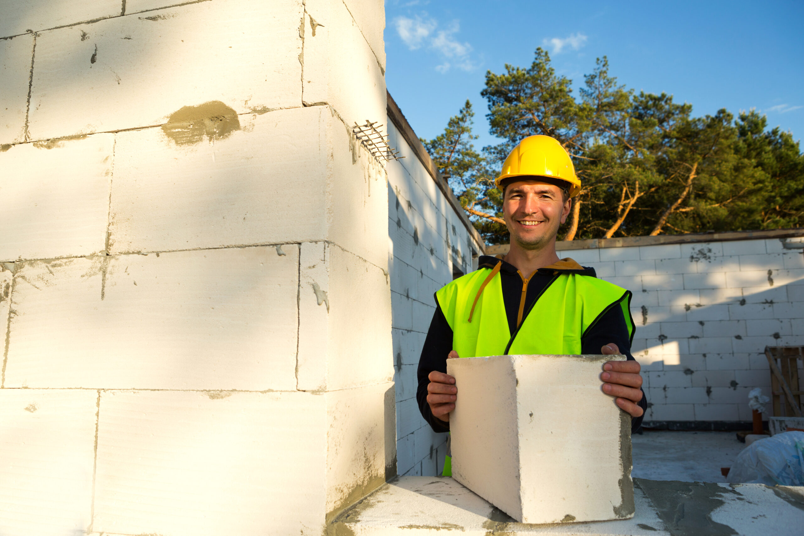 The builder holds a block of cellular concrete in his hands - the masonry of the walls of the house. Construction workers in protective clothing-a hardhat and a reflective vest.